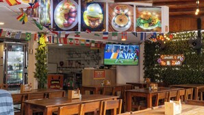 Interior de restaurante com mesas e cadeiras de madeira, uma tela de TV e uma parede decorada com bandeiras e um letreiro de neon.