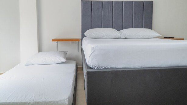 Uma cama de casal com cabeceira cinza, uma cama individual com cabeceira branca e uma mesinha de cabeceira de madeira com um controle remoto.