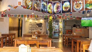Interior de un restaurante con mesas y sillas de madera, una pared decorada con imágenes de comida y un televisor mostrando un partido de fútbol.