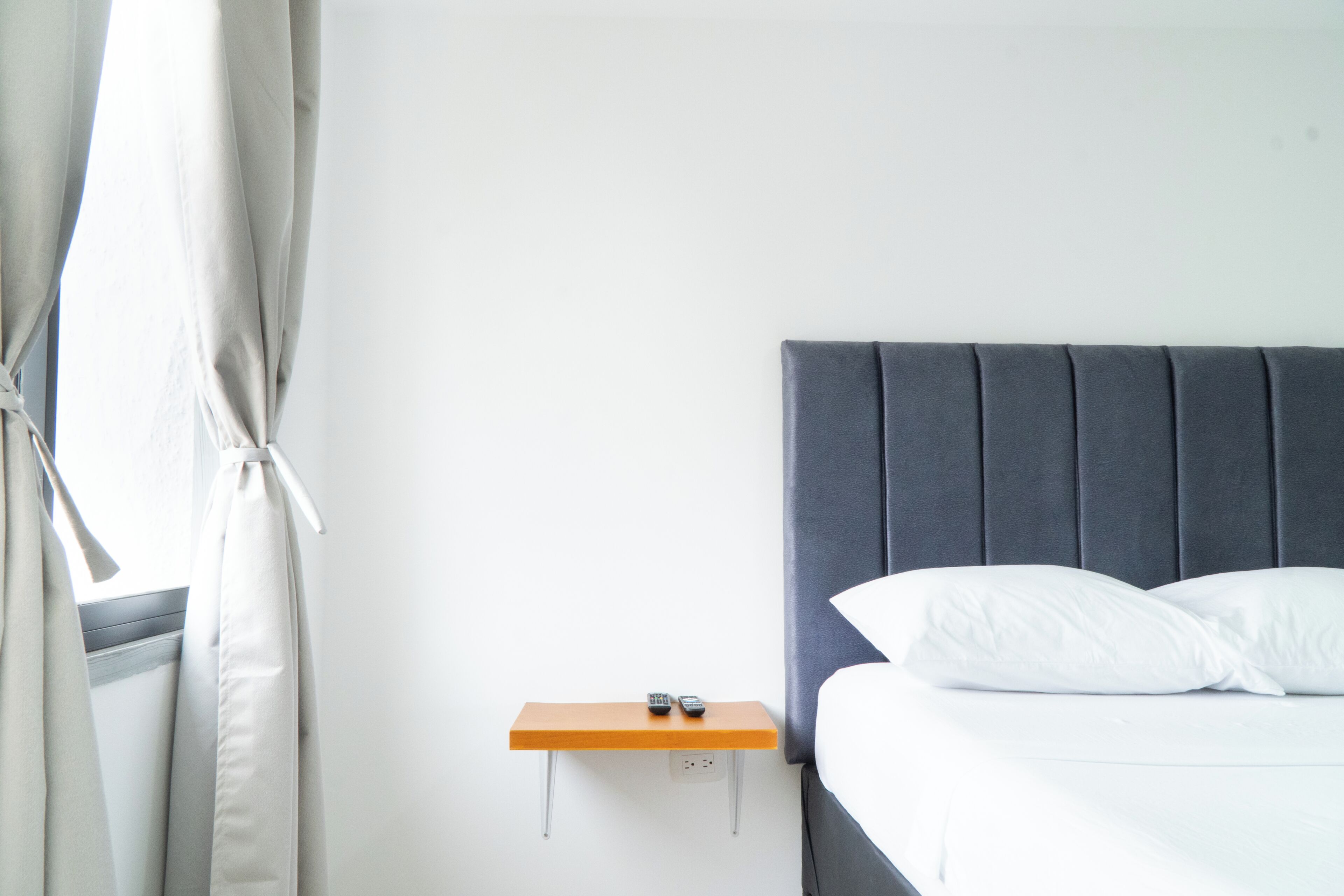A hotel room with a bed, a bedside table with a remote control, and a window with curtains.