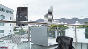 Un balcón con una mesa de vidrio, una computadora portátil y una silla, con vista a un paisaje urbano que incluye edificios y montañas.