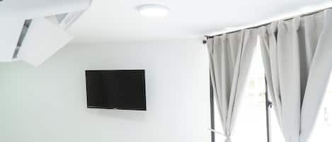 A room with a mounted flat-screen TV on the wall, a window with curtains, and a ceiling-mounted air conditioning unit.