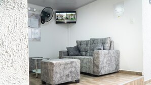 Uma sala de estar moderna com um sofá e um banquinho cinzas, uma pequena mesa e uma TV na parede.