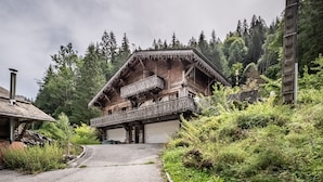 A traditional wooden chalet with a balcony, set in a forested area with a gravel path leading to the entrance.