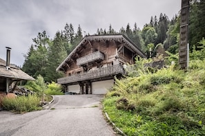 A traditional wooden chalet with a balcony, set in a forested area with a gravel path leading to the entrance.