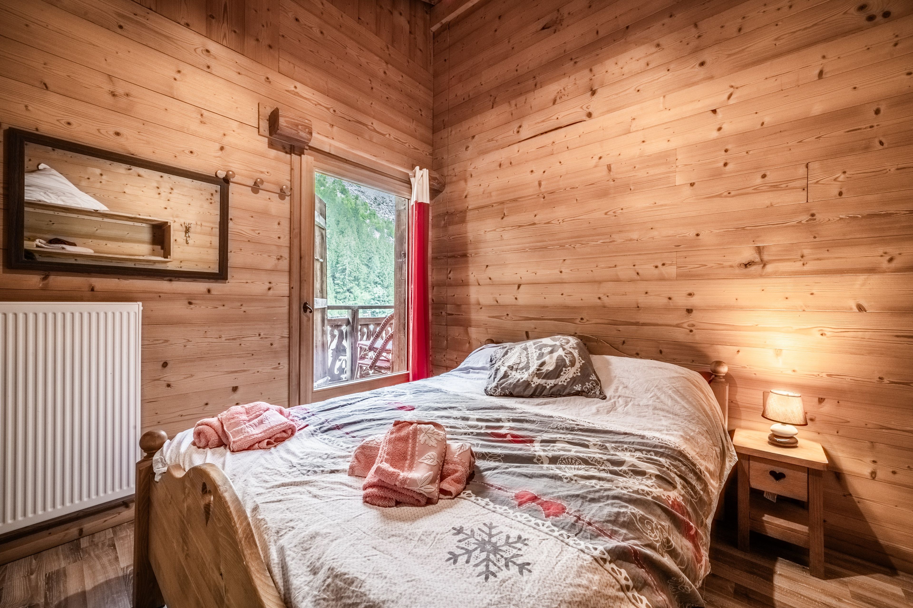 Une chambre dans un chalet en bois, avec un lit, une table de chevet, une fenêtre donnant sur la verdure et un radiateur.