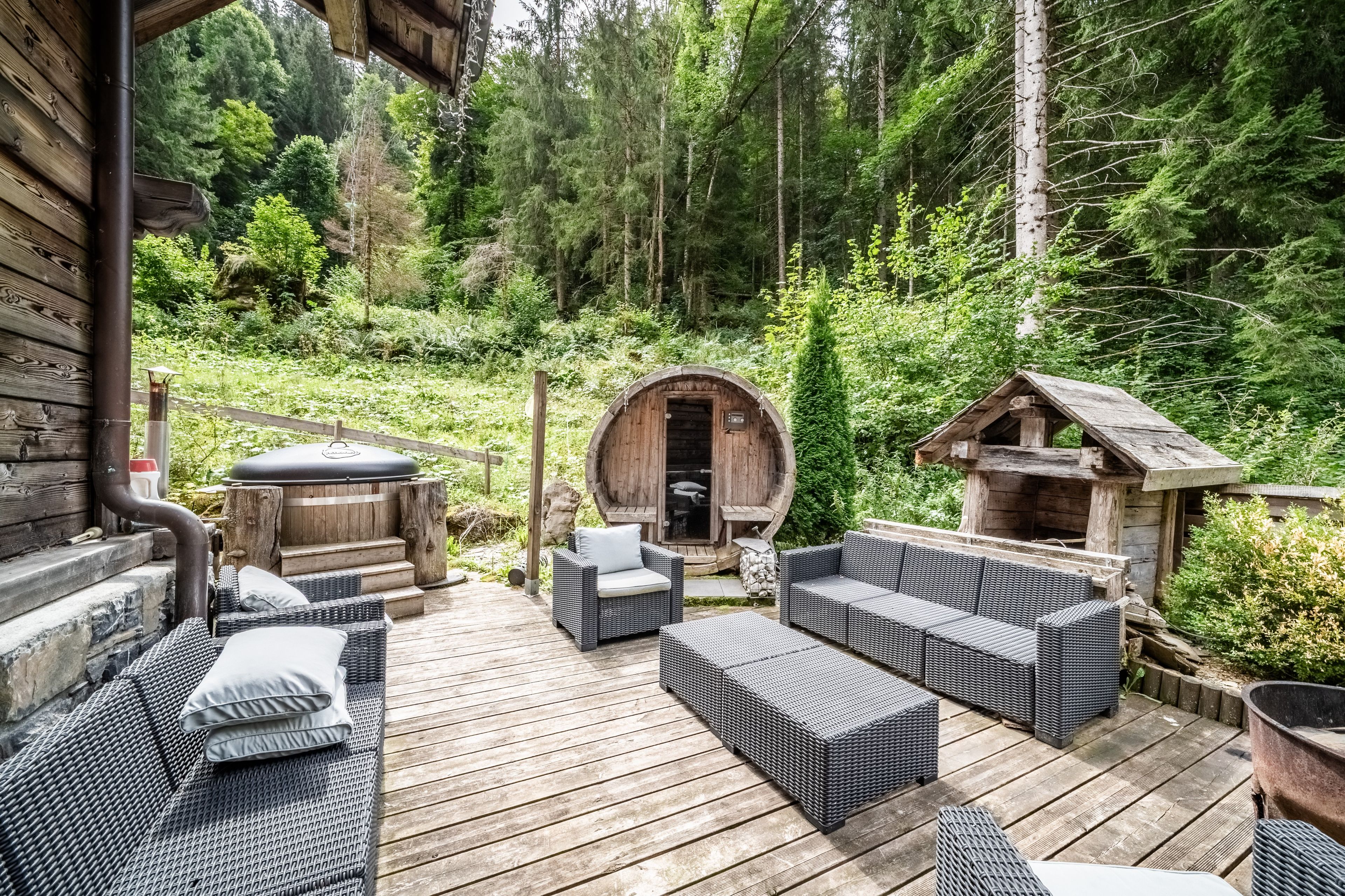 Una terraza de madera con mobiliario de mimbre, un jacuzzi y una estructura con forma de barril en un área boscosa.