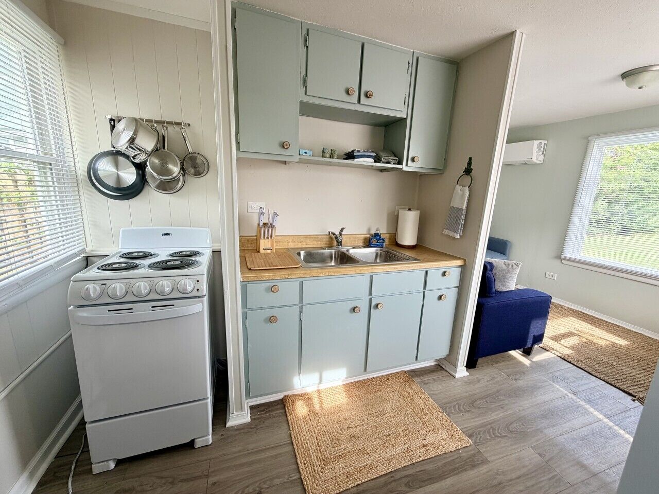 A compact kitchen with a stove, sink, and cabinets, a small seating area, and a window with blinds.