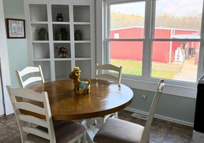 Un comedor con una mesa redonda de madera, sillas blancas y un perro sobre la mesa. Se ve una ventana con vistas a un edificio rojo en el exterior.