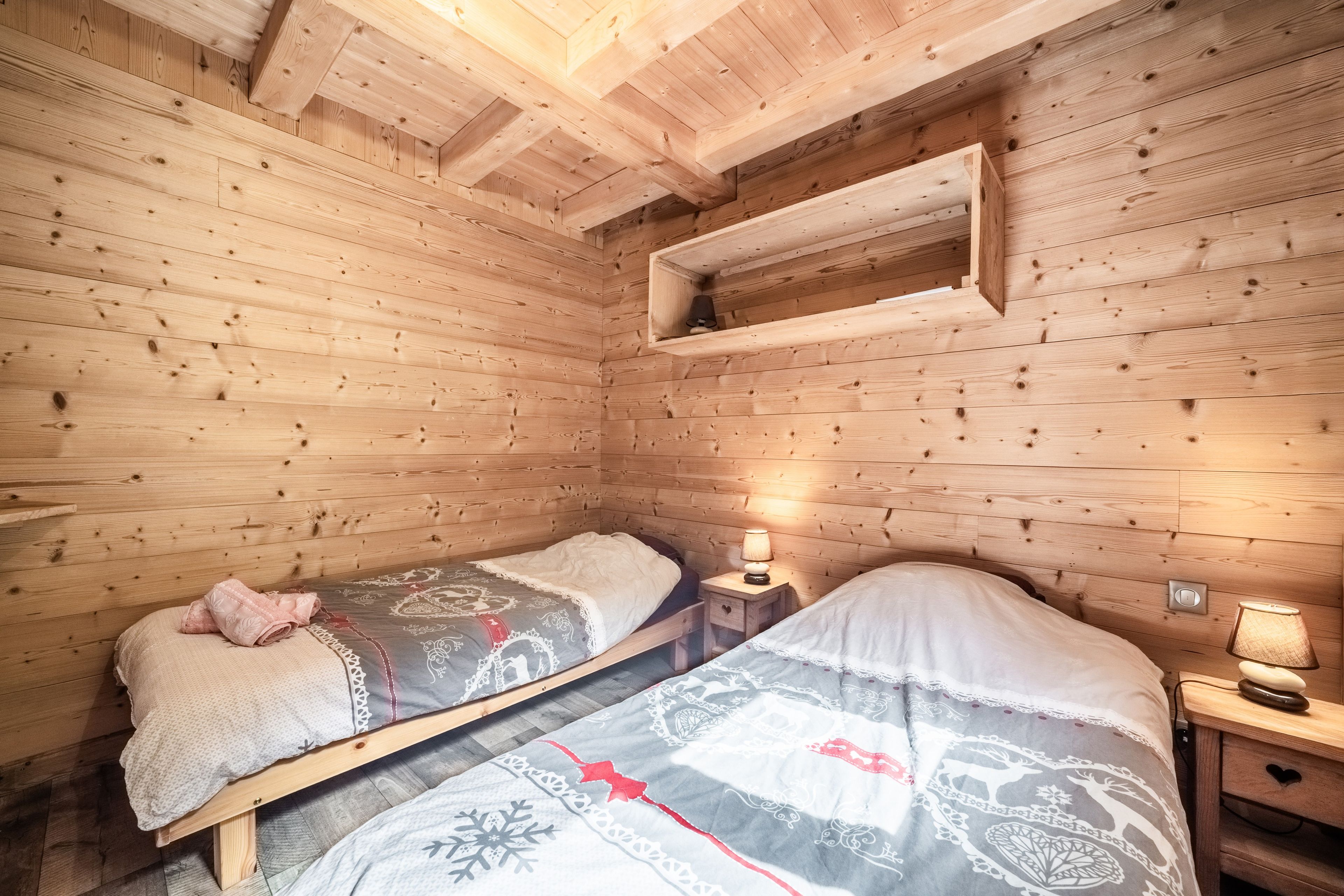 A wooden cabin room with two beds, a shelf, and a nightstand with a lamp.