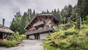 Een klassiek houten chalet met balkon, gelegen in een bebost gebied met een grindpad dat naar de ingang leidt.