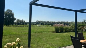 Een overdekt buitengebied met een tafel, stoelen, een gazon en bomen op de achtergrond.
