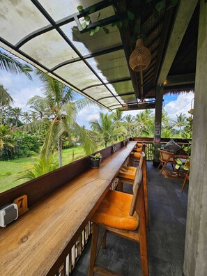 A wooden bar with orange cushioned stools, a thatched roof, and a view of palm trees. - Bali View Ubud Hotel (Gianyar)