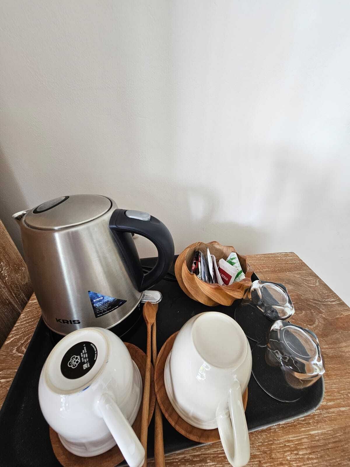 Una tetera eléctrica KRIS de acero inoxidable, dos tazas blancas con tapa, una cuchara de madera, un cuenco de madera pequeño con bolsitas de té y una tetera de vidrio sobre una bandeja negra.