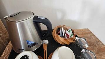 Una tetera eléctrica KRIS de acero inoxidable, dos tazas blancas con tapa, una cuchara de madera, un cuenco de madera pequeño con bolsitas de té y una tetera de vidrio sobre una bandeja negra.