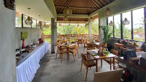 A restaurant with a thatched roof, wooden furniture, and large windows overlooking greenery.