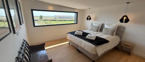 Ein Schlafzimmer mit großem Fenster, einem Bett mit weißer Bettwäsche, einer Holzbank und zwei Pendelleuchten.