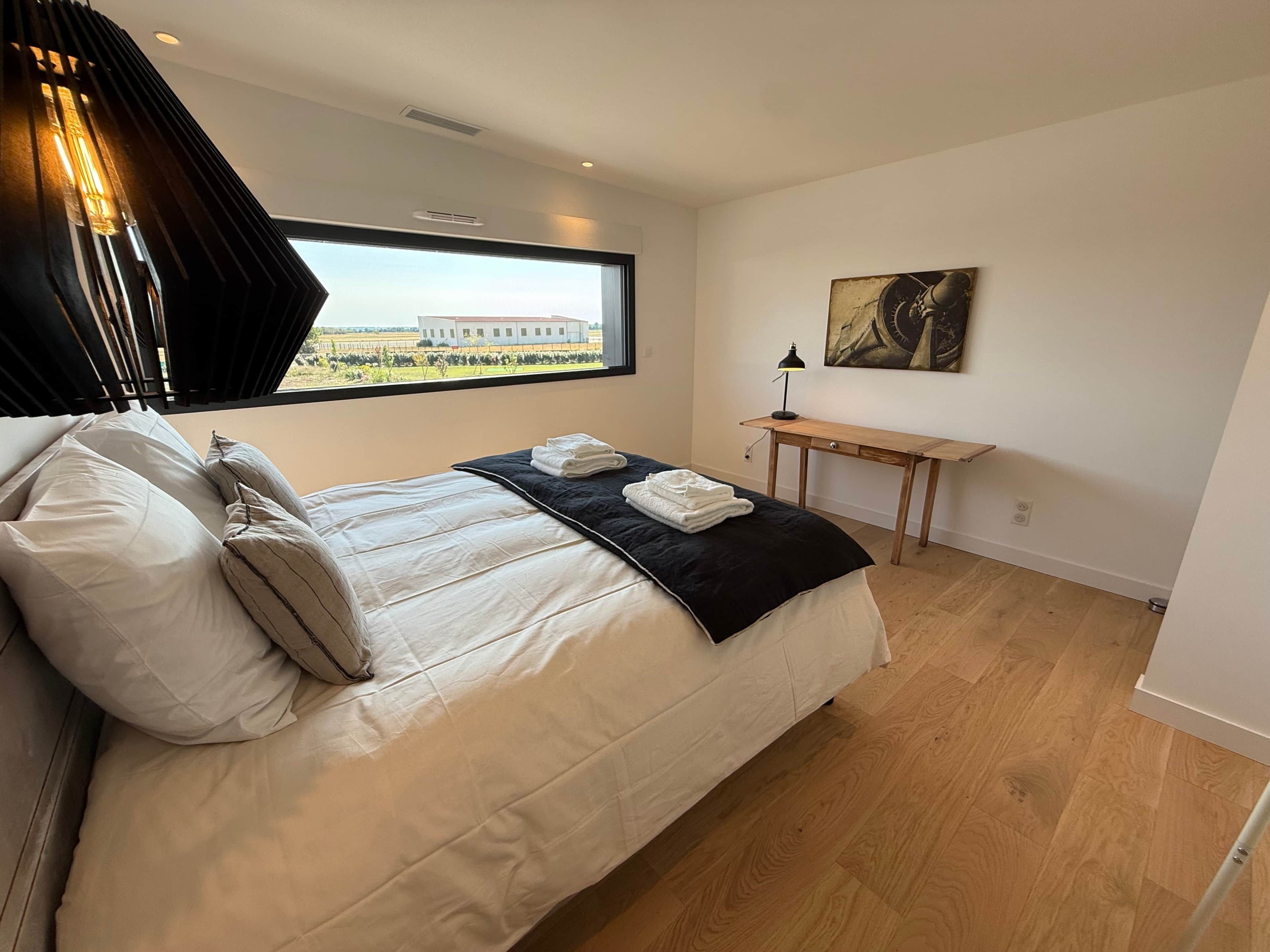 Un dormitorio con cama, almohadas, una mesita de noche con lámpara, una ventana con vista a un edificio y un cuadro en la pared.