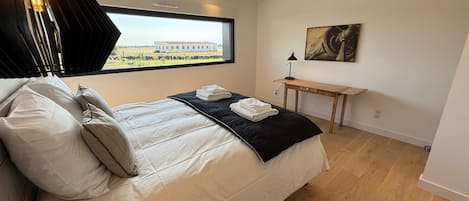 A bedroom with a bed, pillows, a bedside table with a lamp, a window with a view of a building, and a painting on the wall.