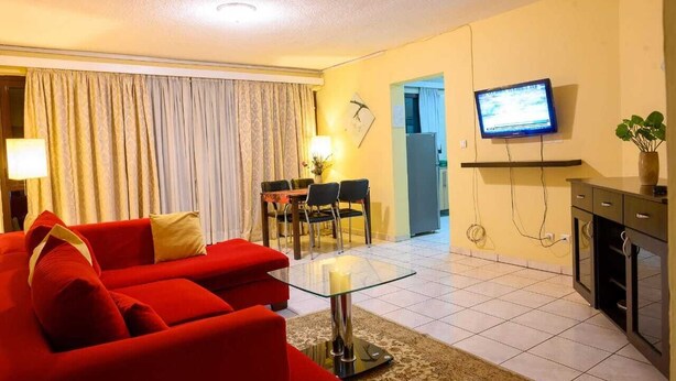 Una sala de estar con un sofá rojo, una mesa de centro de vidrio, un televisor montado en la pared y un comedor con sillas y una mesa.