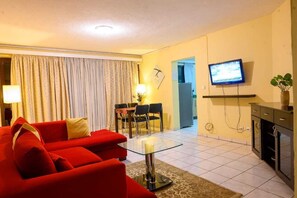 A living room with a red sofa, a glass coffee table, a TV mounted on the wall, and a dining area with chairs and a table. - Residency by La Maison Royale (Kigali)