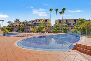 A swimming pool with a tiled edge, surrounded by lounge chairs and palm trees, with hotel buildings in the background. - Vacation Home 'Bungalow Meloneras' with Sea View, Wi-Fi and Air Conditioning (Maspalomas)