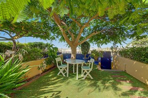 A patio with a table and chairs under a tree, surrounded by greenery and a fence. - Vacation Home 'Bungalow Meloneras' with Sea View, Wi-Fi and Air Conditioning (Maspalomas)