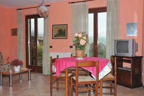 Un área de comedor con una mesa cubierta por un mantel rosa, sillas de madera, una planta en maceta, un televisor en un mueble y una puerta que da al exterior.