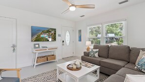 Uma sala de estar com ventilador de teto, televisão, sofá, mesa de centro e vista para o exterior através de grandes janelas.