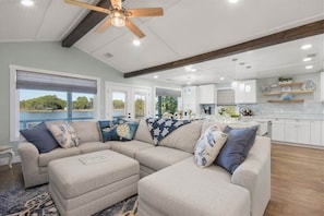 A modern living room with a sectional sofa, ottoman, ceiling fan, and large windows with a view. - The Turtles Nest on Mulat Bay in Avalon Beach (Avalon)