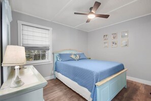 Ein Schlafzimmer mit Bett, Nachttisch, Lampe, Deckenventilator, Fenster mit Jalousien und Wandbildern.