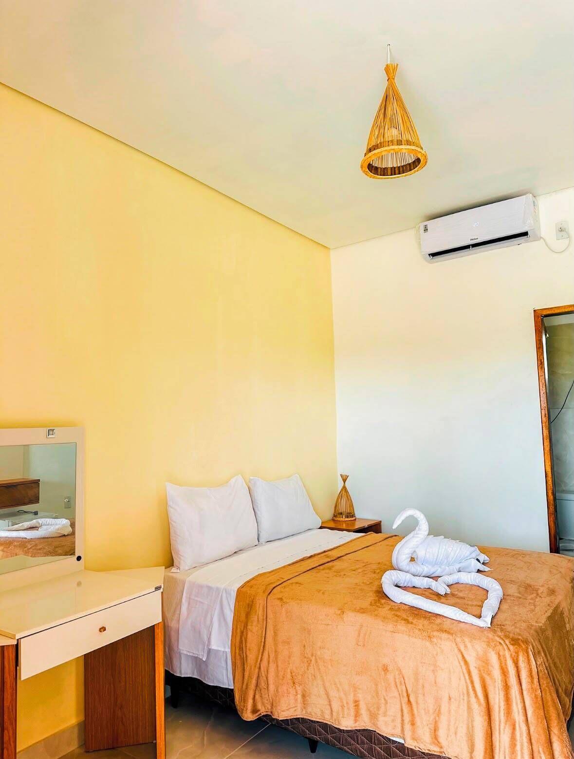 A hotel room with a bed, a bedside table, a mirror, a hanging light fixture, and an air conditioning unit.