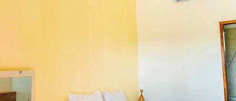A hotel room with a bed, a bedside table, a mirror, a hanging light fixture, and an air conditioning unit.