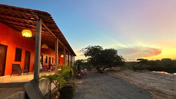 A building with a red facade, a thatched roof, and a paved area with a few plants. - Pousada Caju Sansete  (Barreirinhas)