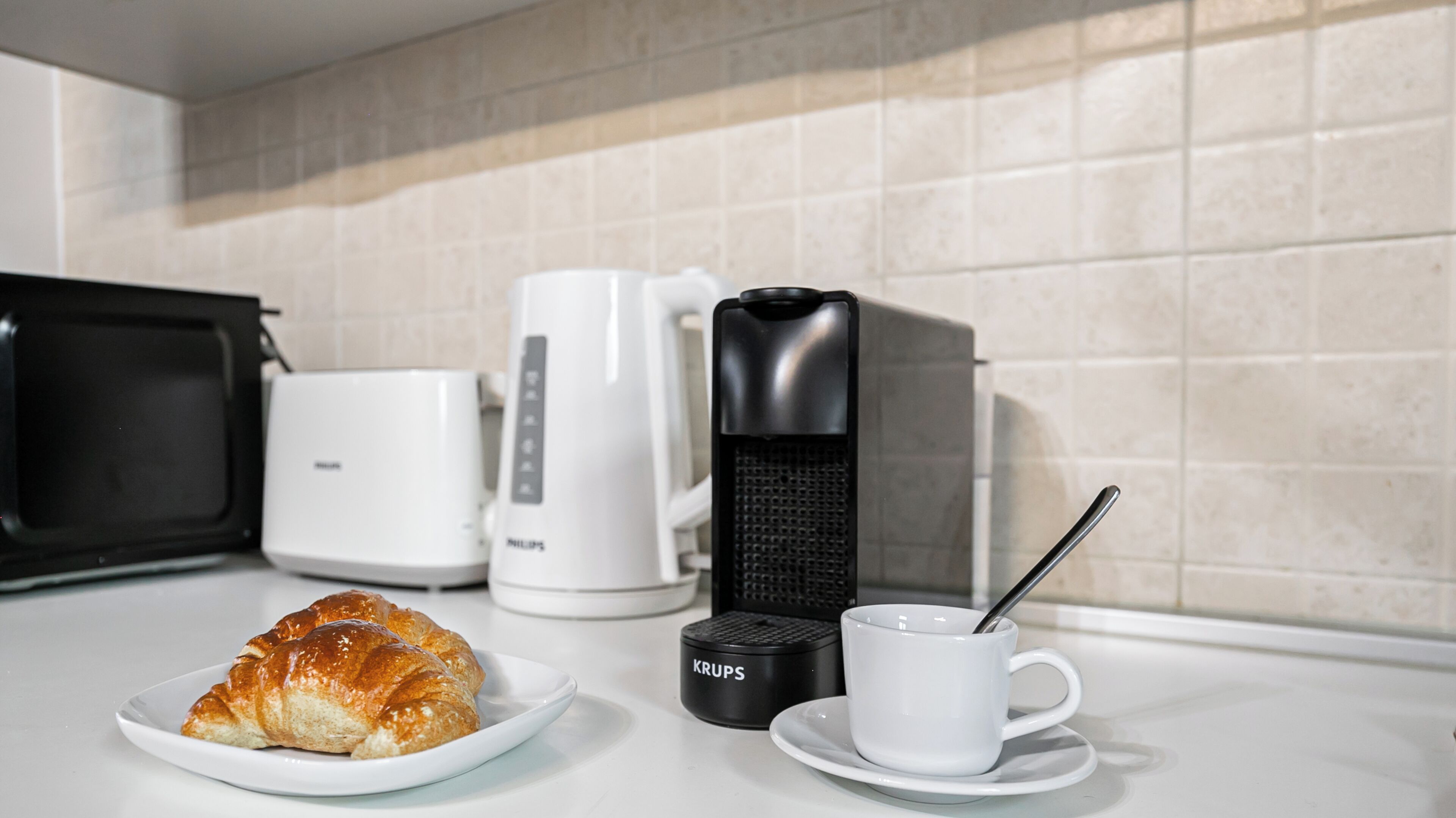 A kitchenette with a microwave, coffee maker, and croissant on a plate.