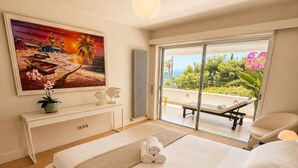 A bedroom with a large bed, a painting of a beach scene, a desk, and a view of the outdoors. - Krabo Villa & Spa (Vouliagmeni)