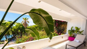 A modern sunroom with a white ceiling, a yellow lounge chair, a white sunbed, and a view of a lush garden and palm trees. - Krabo Villa & Spa (Vouliagmeni)
