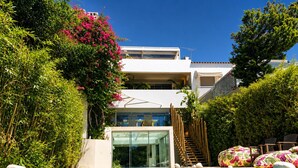 A modern pool area with a clear view of the pool, surrounding greenery, and a multi-level building with a glass entrance. - Krabo Villa & Spa (Vouliagmeni)