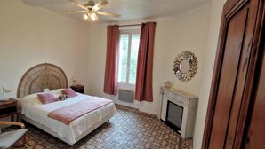 Ein Schlafzimmer mit einem Bett, einem Nachttisch, einem Deckenventilator, einem Fenster mit Vorhängen, einem Kamin und einem Spiegel an der Wand.
