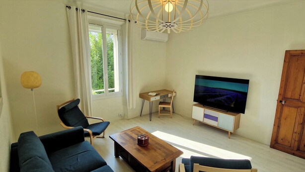 Ein Wohnzimmer mit einer Couch, einem Couchtisch, einem Fernseher auf einem Holzständer, einem Fenster mit Vorhängen und einer Tür.