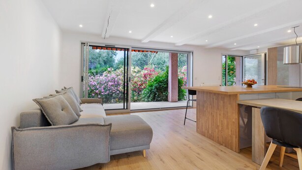 Een moderne woonkamer met een bank, een houten eettafel en een keuken met uitzicht op een tuin door grote schuifdeuren.