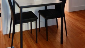 Une pièce avec un plancher en bois, une table blanche avec deux chaises noires, un vase contenant des plantes séchées et un objet blanc posé sur la table.