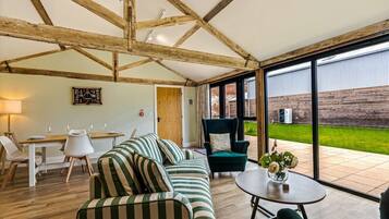 A spacious room with a high ceiling, wooden beams, a dining area, a sofa, a chair, and a view of the outdoors.