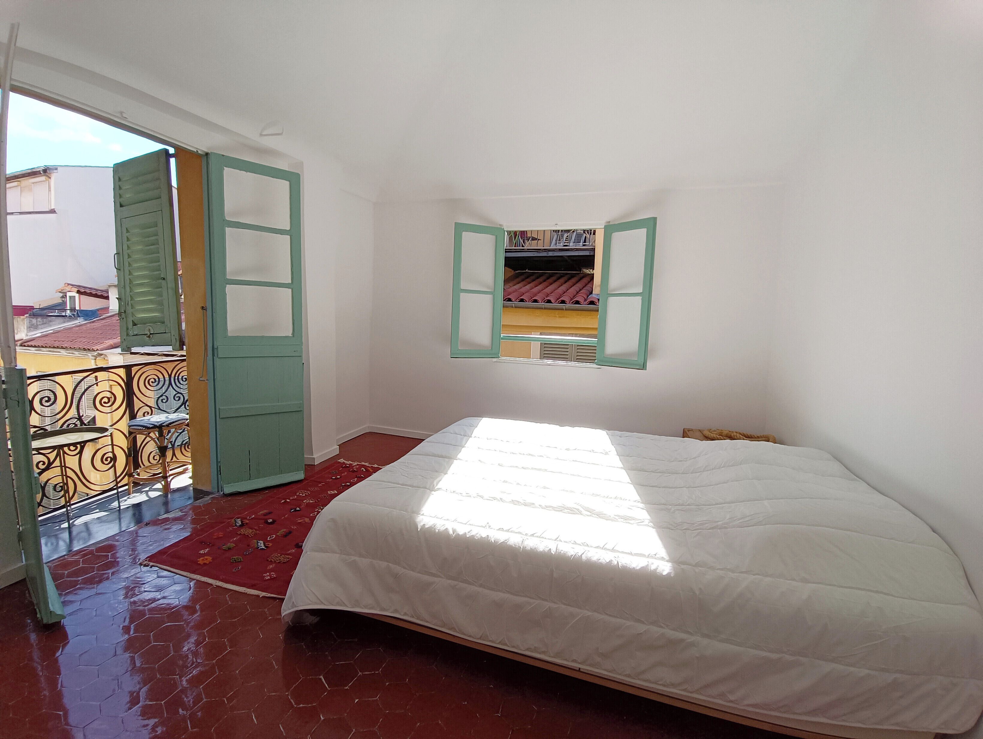 A bedroom with a bed, a window with shutters, a balcony with a railing, and a red patterned rug.