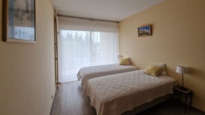A hotel room with two beds, a window with sheer curtains, a painting on the wall, and a small table with a lamp. - Apartment in Mandelieu-la-Napoule (Mandelieu-la-Napoule)