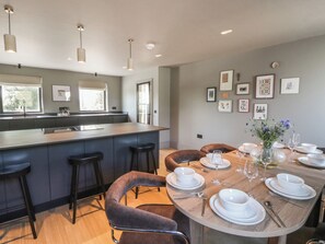 A modern kitchen with a central island, bar stools, and a dining area set for six with white tableware and a vase of flowers. - Brynseion Uchaf (Aberystwyth)