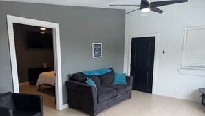 Ein Wohnzimmer mit einem dunkelgrauen Sofa, einem schwarzen Sessel, einem Deckenventilator, einer Tür und einem kleinen Schild an der Wand.