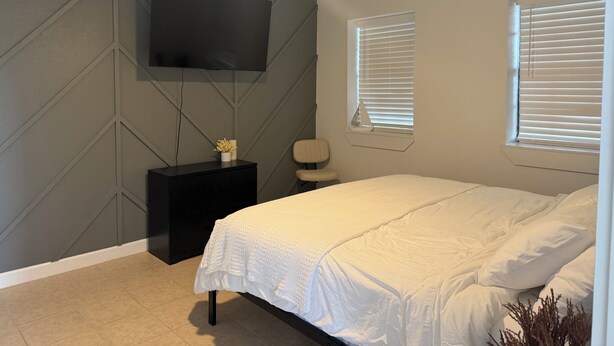 Ein Schlafzimmer mit einem Bett, einem an der Wand montierten Fernseher, einem Deckenventilator und zwei Fenstern mit Jalousien.