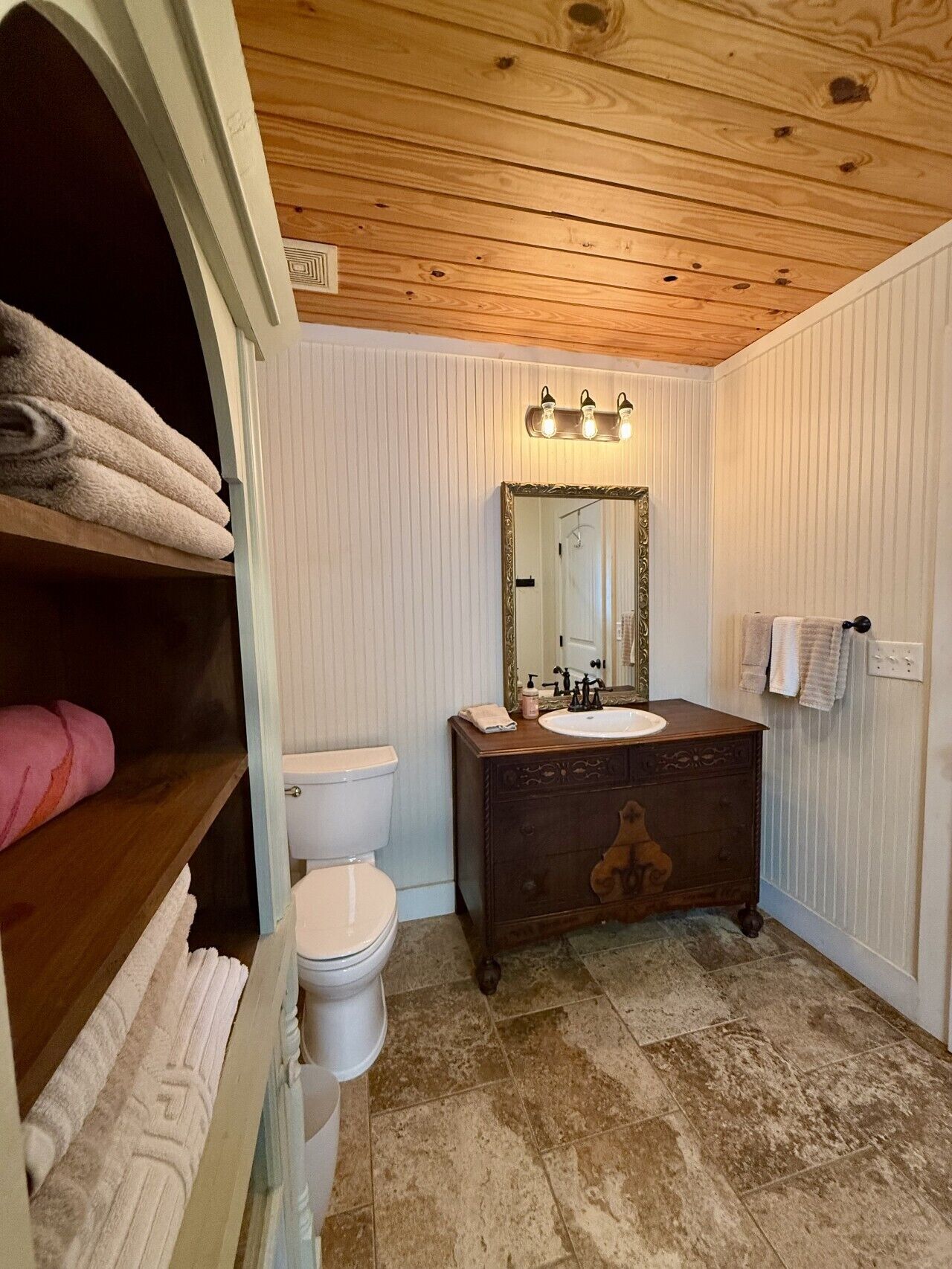 A bathroom with a wooden vanity, a white toilet, and a mirror with lights.
