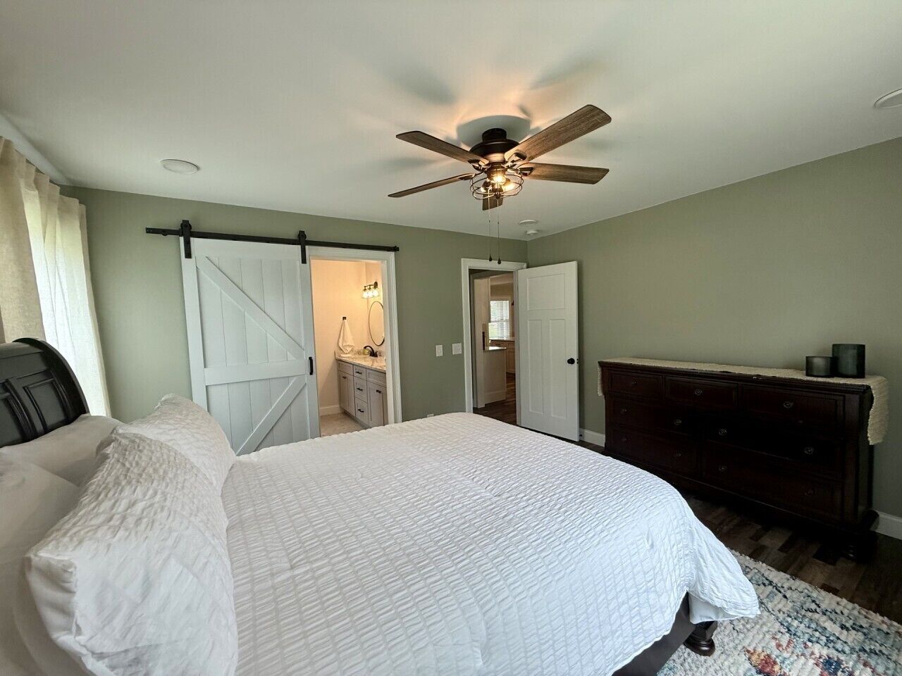Une chambre à coucher comprenant un lit, une commode, un ventilateur de plafond et une porte donnant sur une autre pièce.